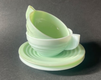 May include: A pale mint green glass teacup and saucer set featuring a distinct ribbed or swirled pattern on the exterior of the cup and the surface of the saucer. The cup has a small handle and a smooth interior finish, crafted from translucent glass.