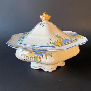 May include: A white ceramic serving dish with a lid, featuring a floral design in yellow, blue, and green. The dish has a scalloped edge and a small, decorative handle on the lid. The base has a unique, curved shape.