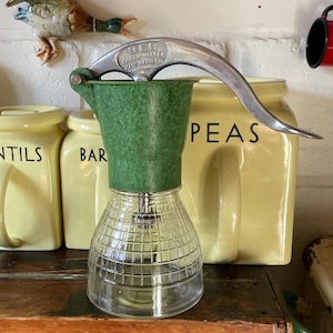 Peut inclure: Un ancien appareil à crème avec un dessus vert et une base en verre transparent. La poignée en métal porte la mention "BEL CREAMMAKER MADE IN ENGLAND". La base en verre présente un motif quadrillé. L'appareil à crème est devant des bocaux jaunes.