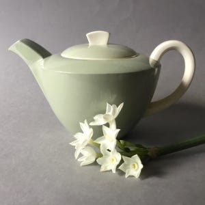 May include: A pale green teapot with a white lid and handle. The teapot is sitting on a gray surface with a sprig of white flowers.