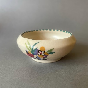 May include: A small, round, cream-coloured ceramic bowl with a subtle crackle glaze finish. The exterior features a hand-painted floral motif in shades of yellow, blue, green, and red, with a decorative teal dashed pattern along the rim.