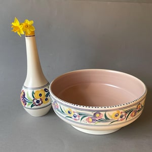 May include: A cream-coloured vase and a bowl with floral designs. The vase has a slender neck and a bulbous base, with yellow daffodils. The bowl has a pink interior and a floral border with yellow, purple, and blue flowers.
