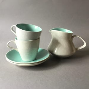 Könnte beinhalten: Ein Set aus drei hellblauen Keramik-Teetassen und Untertassen mit einem passenden hellblauen Keramik-Milchkännchen. Die Tassen und das Milchkännchen haben eine strukturierte, leicht irisierende Glasur.