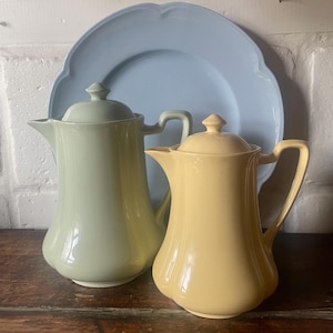 Può includere: Due brocche in ceramica, una verde pallido e una gialla, con coperchi abbinati e manici curvi. Dietro c'è un piatto azzurro smerlato. Le brocche sono su una superficie di legno.