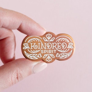 May include: A copper-colored enamel pin with a white design. The pin features a stylized floral design with the words "KINDRED SPIRIT" in a script font.