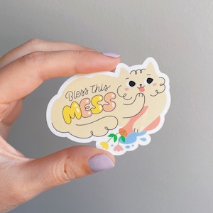 Könnte beinhalten: Ein weißer Aufkleber mit einer Cartoon-Illustration eines flauschigen Hundes mit dem Text "Bless this mess" in gelben und rosa Buchstaben.
