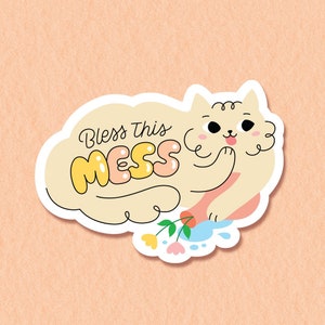 Peut inclure: Un autocollant blanc avec un chat de dessin animé et le texte "Bless this mess" en lettres colorées. Le chat tient une fleur et il y a des fleurs et des gouttelettes d'eau autour de lui.