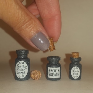 Pode incluir: Três pequenas garrafas de vidro com rolhas de cortiça. As garrafas são pretas com rótulos brancos. Os rótulos dizem "Deadly Nightshade", "Frog's Breath" e "Worm's Wort".