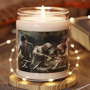 Puede incluir: Una vela encendida en un tarro de cristal con una etiqueta que presenta una ilustraci&oacute;n vintage y el texto "The Infernal Devices". La vela est&aacute; sobre un posavasos de madera, rodeada de luces de hadas.