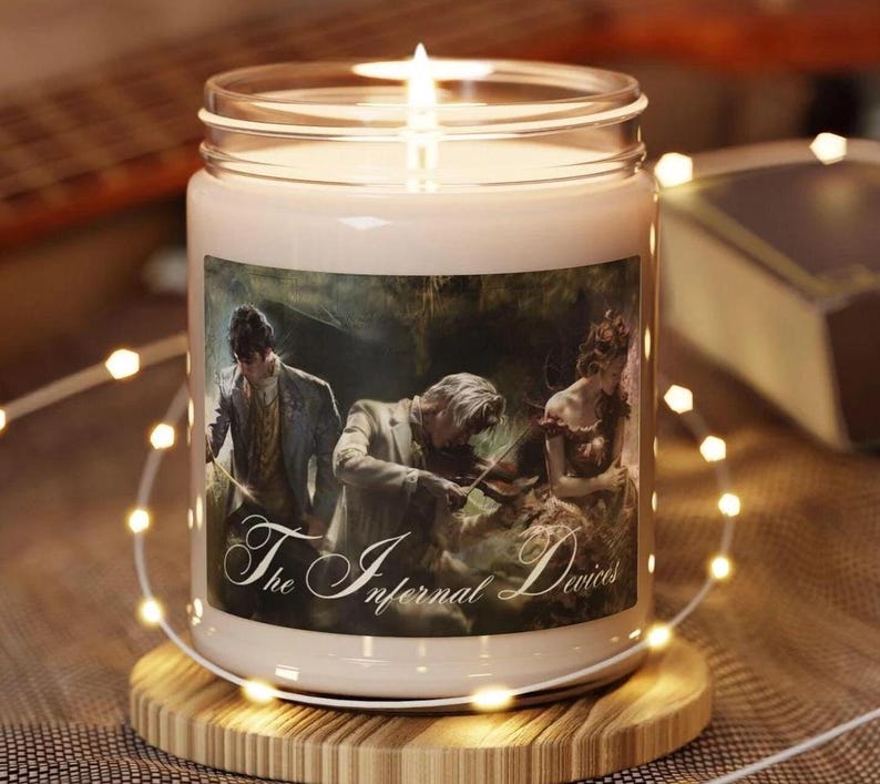 Puede incluir: Una vela encendida en un tarro de cristal con una etiqueta que presenta una ilustraci&oacute;n vintage y el texto "The Infernal Devices". La vela est&aacute; sobre un posavasos de madera, rodeada de luces de hadas.