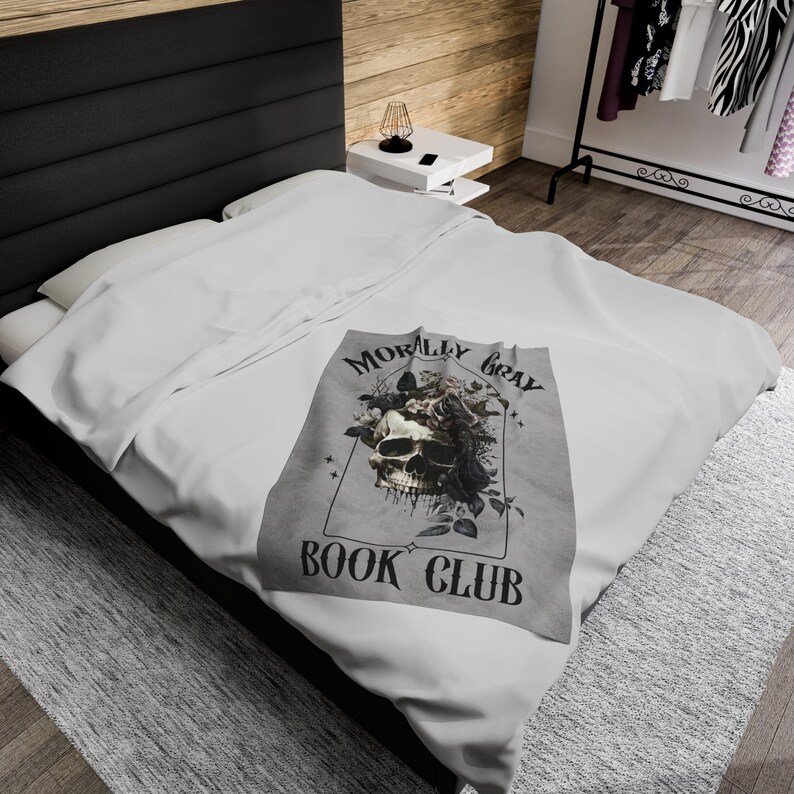May include: A gray blanket with a skull and floral design, with the words "MORALLY GRAY BOOK CLUB" printed on it. The blanket is on a white bed, with a black headboard and a wooden wall in the background.