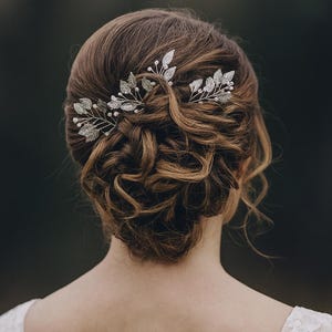 May include: A woman's updo hairstyle with silver leaf hairpins. The hairpins are adorned with small pearls and are placed in the updo. The hair is a light brown color, and the background is a blurred dark green.