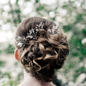 May include: A woman with curly brown hair wearing a silver hair comb with leaf and pearl accents.