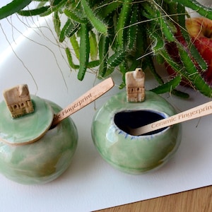 May include: Two round, light green ceramic bowls with lids, each with a small wooden spoon. The lids are topped with a small house-shaped design. The spoons are engraved with "Ceramic Fingerprint". A red apple and green plant are in the background.