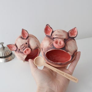 May include: Two ceramic pig-shaped bowls with open mouths and pink interiors. The pigs have pink ears and snouts. A small wooden spoon with the text "Ceramic Fingerprint" is held near the bowls. A silver bell is in the background.