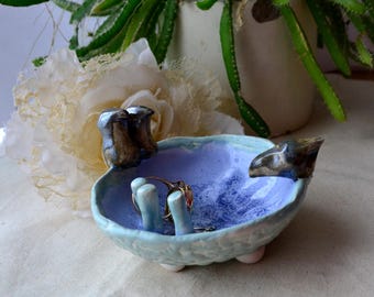 Könnte beinhalten: Eine hellblaue Keramikschale mit strukturierter Oberfläche und drei Füßen. Die Schale hat ein violettes Inneres und verfügt über zwei kleine Keramikvögel und einen Ringhalter.