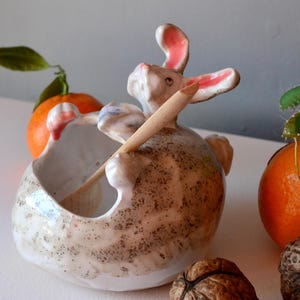 Puede incluir: Un tazón de cerámica con forma de conejo que sostiene una cuchara de madera. El tazón está salpicado de esmalte marrón y blanco. El conejo tiene orejas rosas y un cuerpo blanco.