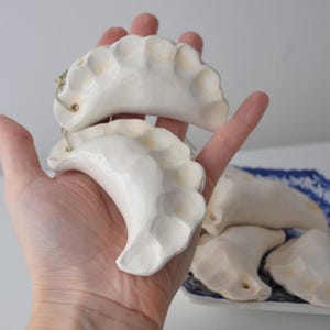 May include: Off-white ceramic pierogi ornaments with yellow highlights. Two are held in a hand, with more on a blue and white patterned plate. These unique ornaments add a touch of whimsy to any holiday decor. Each pierogi is approximately 7 cm long.