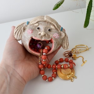 May include: A ceramic face-shaped trinket dish with an open mouth, holding a ring and a coral bead necklace. Other jewellery, including rings and a gold hat pin, are next to the dish. The face has pink cheeks and blue hair ties.