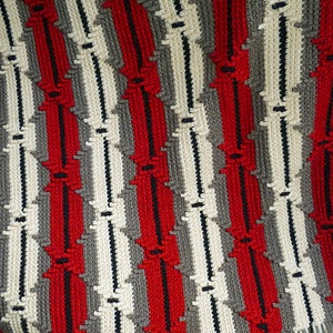 Pode incluir: Um cobertor feito à mão com um padrão repetido de listras verticais vermelhas, brancas, cinzas e pretas. O cobertor tem uma borda com franjas com fios vermelhos e brancos. O design é uma série de formas de diamante conectadas.