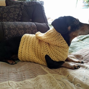 May include: A black and tan dachshund wearing a hooded, yellow crocheted sweater. The dog is lying on a patterned bedspread. The sweater has a textured, open weave design.