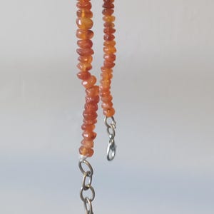 Peut inclure: Un collier de perles orange avec une chaîne argentée. Les petites perles à facettes créent un effet scintillant. Le collier est suspendu à une barre argentée sur fond blanc.