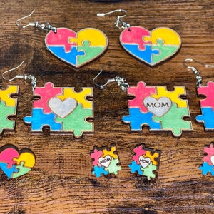 May include: A set of six pairs of wooden puzzle piece earrings. The earrings are painted in various colors, including pink, blue, green, and yellow. Some of the earrings feature a heart shape in the center, while others have the word "MOM" inscribed.