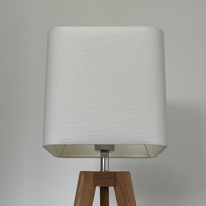 May include: A contemporary floor lamp featuring a rectangular, off-white lampshade. The lampshade sits on a chrome stem, supported by a wooden tripod base. The wood has a natural finish.