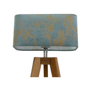 May include: A teal rectangular lampshade with gold detailing sits atop a chrome stem and wooden tripod base. The lamp is designed for indoor use. The base is made of wood.