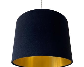 May include: A black lampshade with a gold interior. The lampshade is cylindrical with a slightly tapered shape. It is suspended from a thin wire, and the interior has a shiny gold finish.