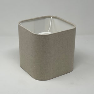 May include: A square lampshade with a neutral beige fabric exterior and a white interior. The lampshade has a rounded square shape and a white metal frame. The lampshade is designed to diffuse light.