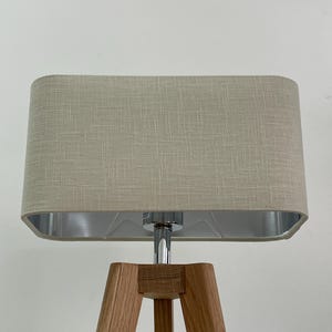 May include: A beige linen lampshade with a silver lining sits atop a wooden tripod lamp base.