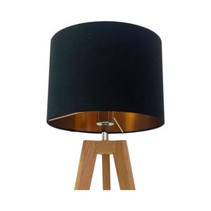 May include: A contemporary table lamp featuring a black lampshade with a gold interior. It has a wooden tripod base and a silver metal stem. The lampshade is cylindrical in shape.