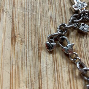 Puede incluir: Pulsera plateada con dijes de coraz&oacute;n y cruz. La pulsera presenta una cadena de cuerda trenzada y un cierre de mosquet&oacute;n. Los dijes incluyen un coraz&oacute;n con un dise&ntilde;o grabado y dijes en forma de cruz con piedras transparentes.