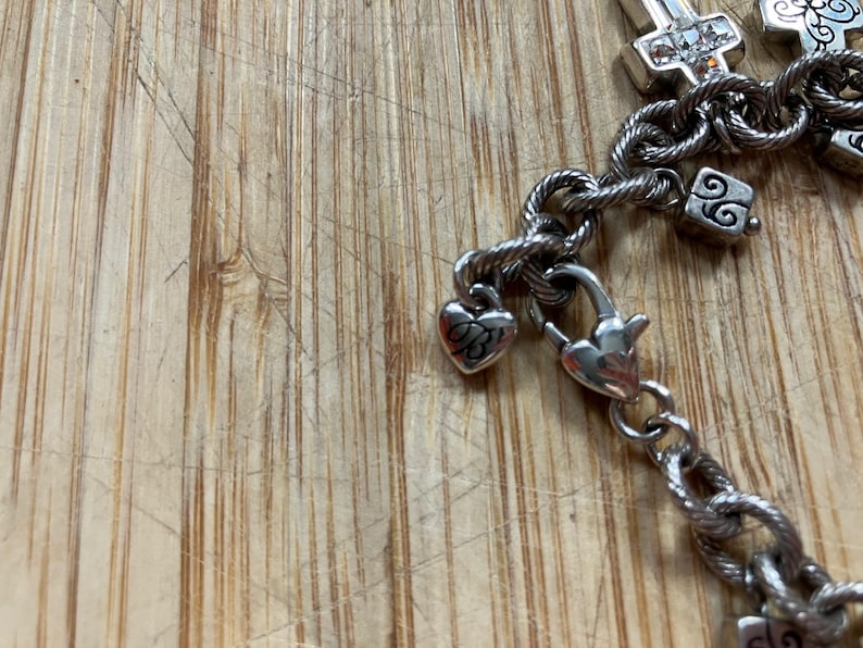 Puede incluir: Pulsera plateada con dijes de coraz&oacute;n y cruz. La pulsera presenta una cadena de cuerda trenzada y un cierre de mosquet&oacute;n. Los dijes incluyen un coraz&oacute;n con un dise&ntilde;o grabado y dijes en forma de cruz con piedras transparentes.