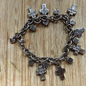 Puede incluir: Pulsera de abalorios plateada con abalorios en forma de cruz, algunos con detalles de cristal transparente. La pulsera tiene una cadena texturizada y un cierre en forma de coraz&oacute;n. Los abalorios incluyen volutas decorativas.