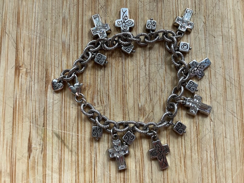 Puede incluir: Pulsera de abalorios plateada con abalorios en forma de cruz, algunos con detalles de cristal transparente. La pulsera tiene una cadena texturizada y un cierre en forma de coraz&oacute;n. Los abalorios incluyen volutas decorativas.