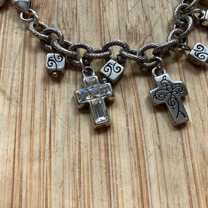 Puede incluir: Pulsera de abalorios plateada con un dise&ntilde;o de cadena retorcida. La pulsera presenta varios dijes de cruz, algunos adornados con cristales transparentes, y peque&ntilde;os dijes cuadrados con remolinos decorativos. El fondo es una superficie de madera marr&oacute;n claro.