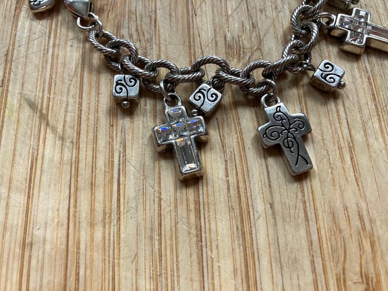 Puede incluir: Pulsera de abalorios plateada con un dise&ntilde;o de cadena retorcida. La pulsera presenta varios dijes de cruz, algunos adornados con cristales transparentes, y peque&ntilde;os dijes cuadrados con remolinos decorativos. El fondo es una superficie de madera marr&oacute;n claro.