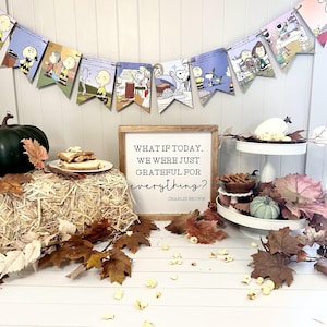 May include: A Thanksgiving-themed table setting with a wooden sign that reads "What if today, we were just grateful for everything? Charlie Brown". The table is decorated with fall leaves, popcorn, and a tiered serving tray with snacks.