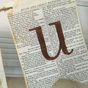 Op de afbeelding: Een bruine papieren banner met de letter "U" gedrukt in bruine verf. De banner is gemaakt van een pagina uit een boek met tekst uit Shakespeares "Veel drukte voor niets".