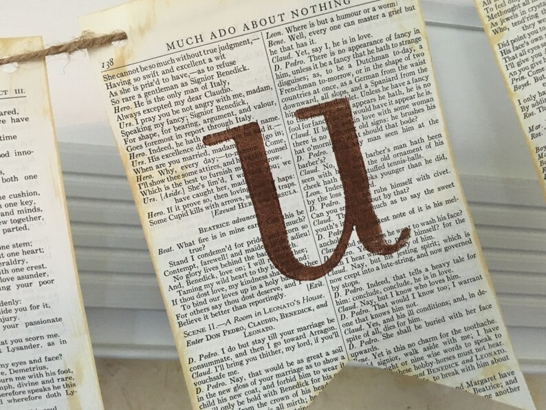 Op de afbeelding: Een bruine papieren banner met de letter "U" gedrukt in bruine verf. De banner is gemaakt van een pagina uit een boek met tekst uit Shakespeares "Veel drukte voor niets".