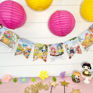 May include: A white and blue banner with illustrations from the Disney movie Snow White and the Seven Dwarfs. The banner is strung between two pink paper lanterns and two yellow paper lanterns. The banner is hanging on a white wall. There are also some small decorations on a pink table in the foreground.