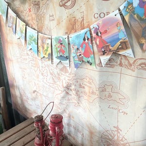 K&ouml;nnte beinhalten: Eine Piratenkarte im Vintage-Stil mit einem Kompass und einer Girlande aus bunten Flaggen mit Figuren aus Peter Pan. Die Flaggen zeigen Captain Hook, Peter Pan und Tinkerbell. Die Karte ist auf braunem Papier mit einem verwitterten Look gedruckt.
