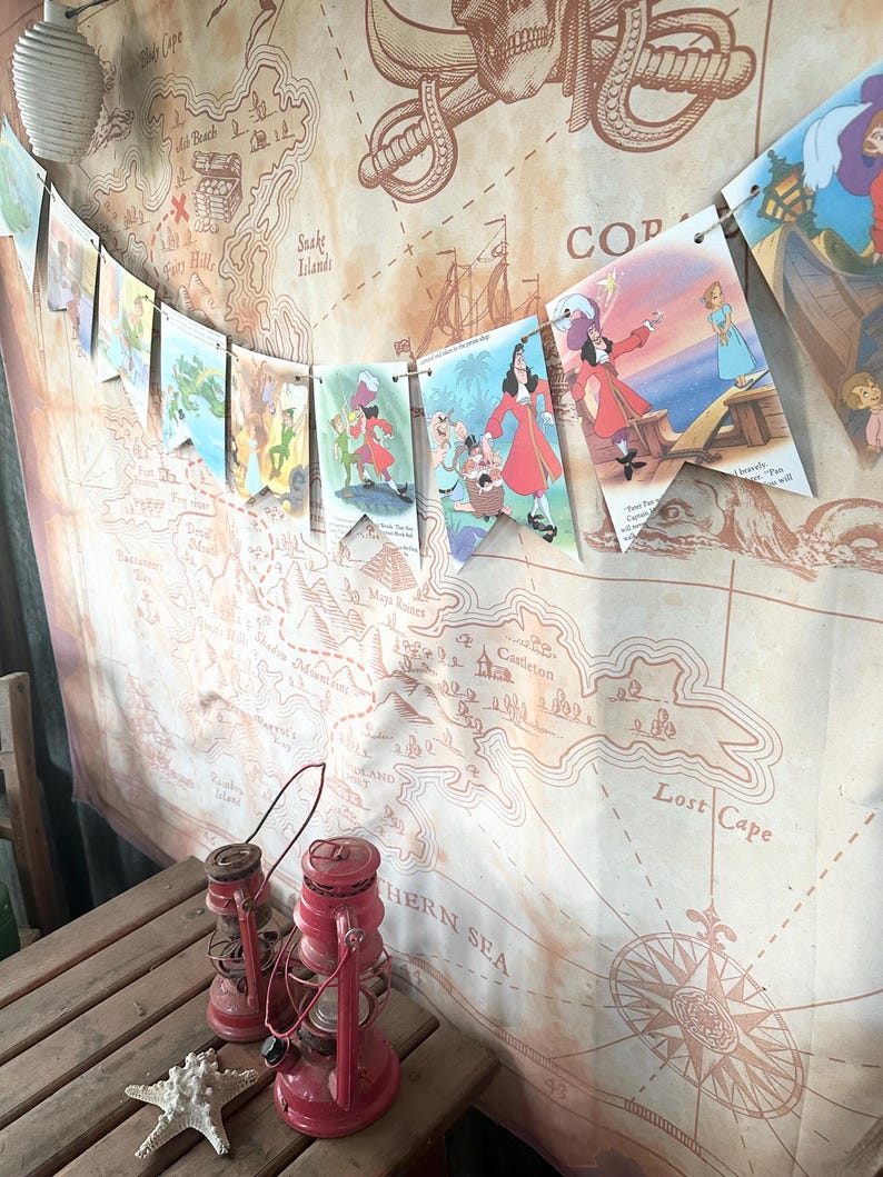 K&ouml;nnte beinhalten: Eine Piratenkarte im Vintage-Stil mit einem Kompass und einer Girlande aus bunten Flaggen mit Figuren aus Peter Pan. Die Flaggen zeigen Captain Hook, Peter Pan und Tinkerbell. Die Karte ist auf braunem Papier mit einem verwitterten Look gedruckt.