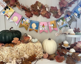 May include: A Halloween-themed banner with Peanuts characters, featuring a pink, yellow, and blue color scheme. The banner is decorated with fall leaves and pumpkins.