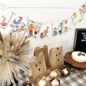 May include: A festive party scene with a Paddington Bear theme. Includes brown paper bear-shaped treat bags, cupcakes, a cake with bear-shaped decorations, and a banner with illustrations. A sign reads "PLEASE FEED THE BEARS."