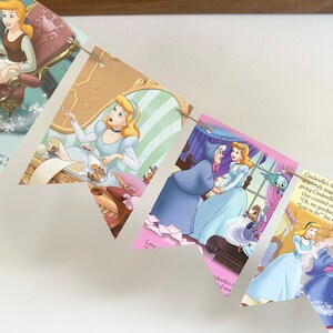 May include: A decorative banner featuring illustrations from the Cinderella story. The pennants display scenes of Cinderella in her blue gown, with text from the story. The banner is strung on twine and is set against a white background.