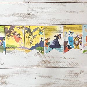 May include: A series of colorful, illustrated pennant banners featuring scenes from The Wizard of Oz. The banners depict characters like Dorothy, the Wicked Witch, the Tin Man, and the Lion. The illustrations have a vintage, storybook style, with a white wooden background.