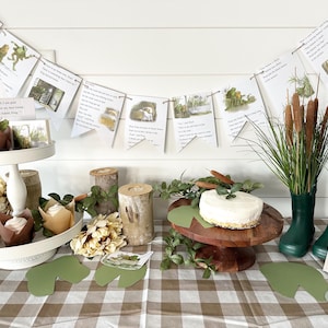 May include: A Frog and Toad themed party table setting with a white and brown checkered tablecloth. The table is decorated with green leaves, brown logs, and a two-tiered white cake stand with brown cupcake liners. There is a white cake with green frosting on a wooden cake stand. There is a banner with illustrations of Frog and Toad hanging on the wall. The text on the banner reads "Dear Toad, I am glad you are my best friend. Your best friend, Frog."